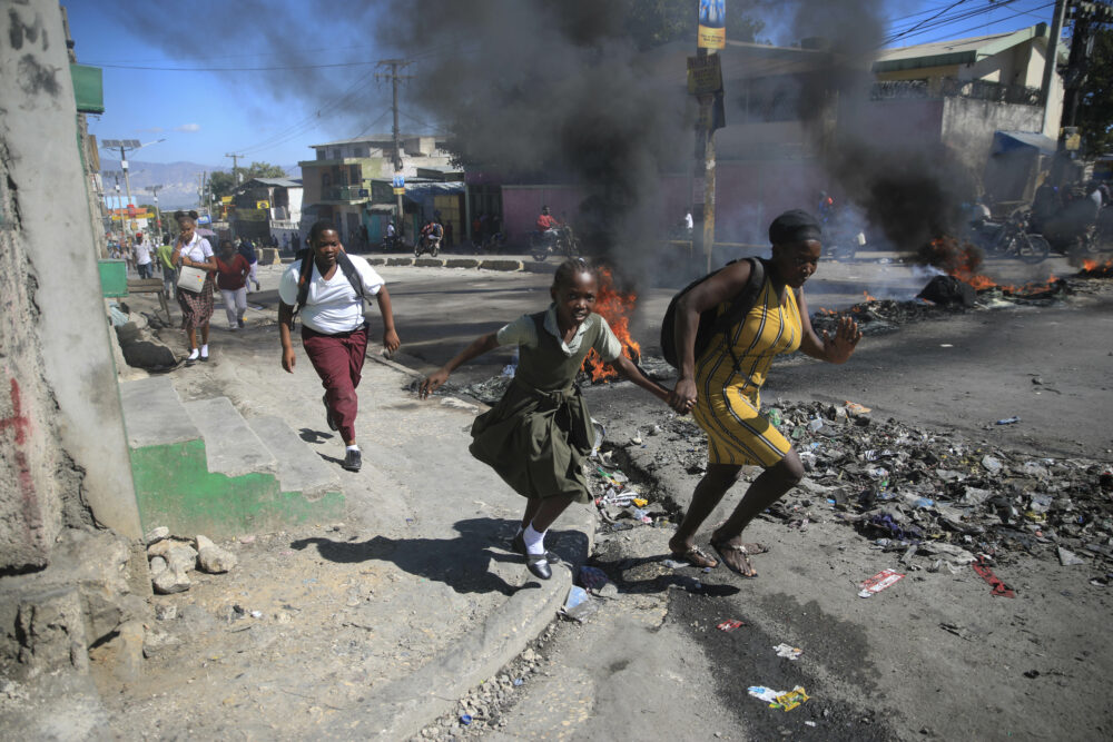 Archivo - Una mujer y su hija pasan corriendo frente a una barricada levantada por la policía en protesta por el mal gobierno policial en Puerto Príncipe (AP Foto/Odelyn Joseph, Archivo)